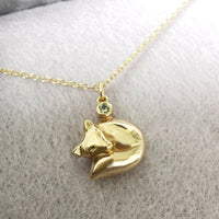 A gold necklace with a small fox pendant resting on a soft, light gray textured fabric background. The pendant features a tiny green gemstone accent at the top.