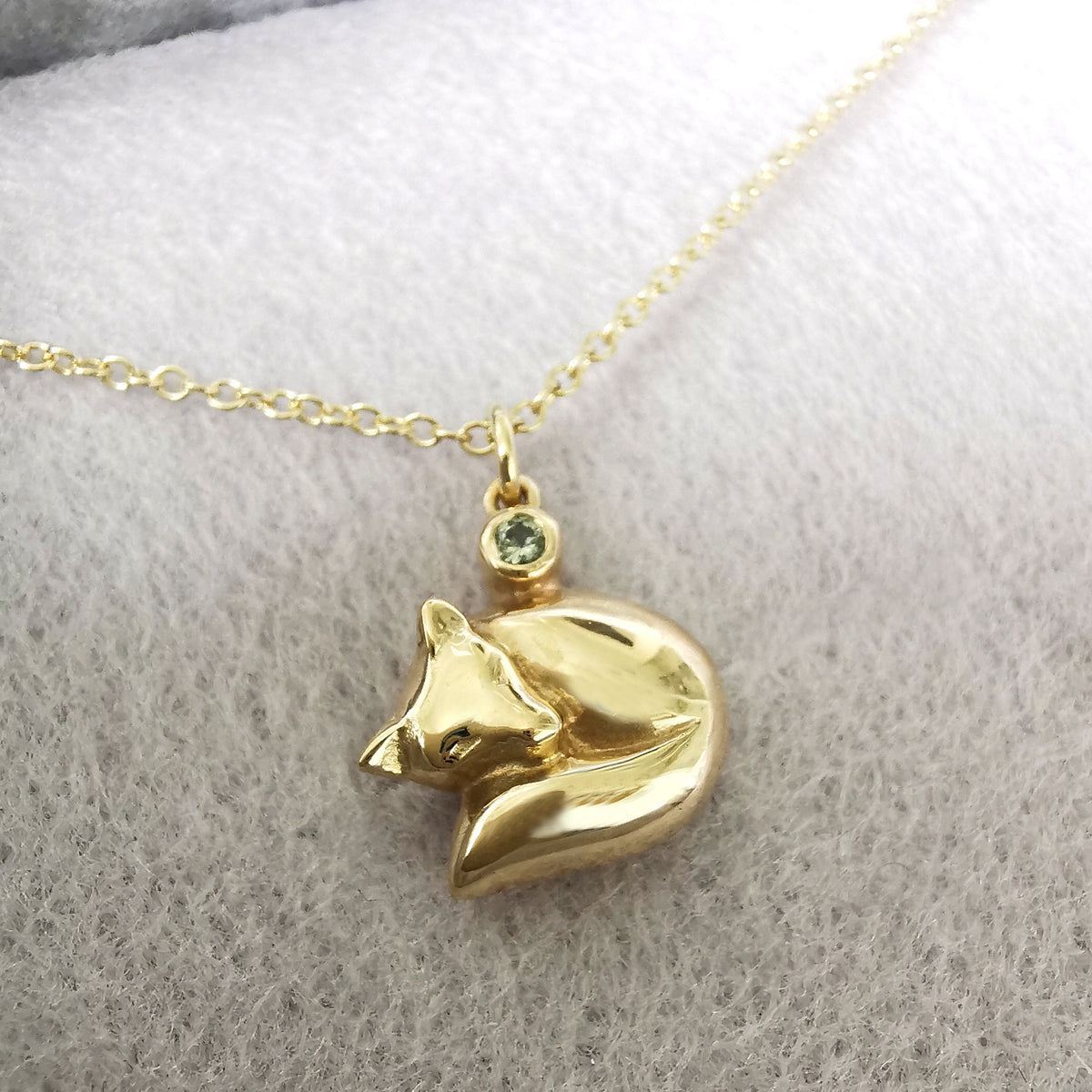 A gold necklace with a small fox pendant resting on a soft, light gray textured fabric background. The pendant features a tiny green gemstone accent at the top.